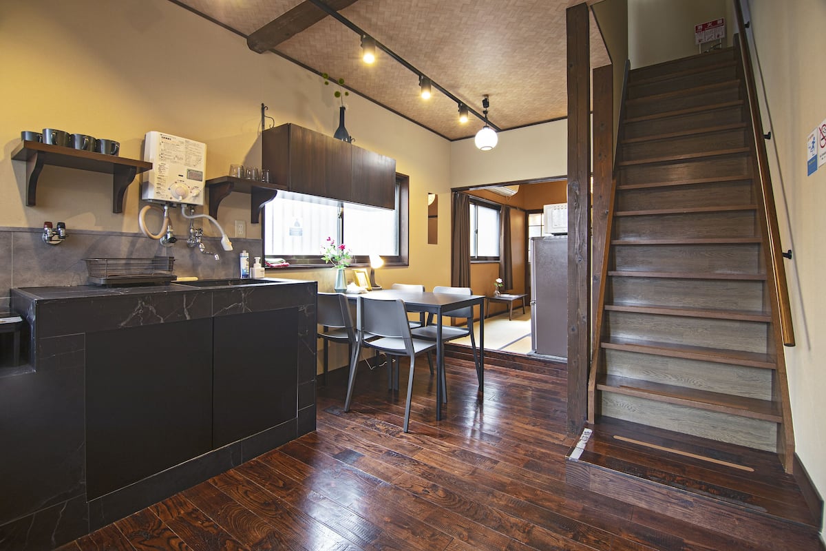A kitchen with dark cabinetry, a dining area with a table and chairs, and a wooden staircase.