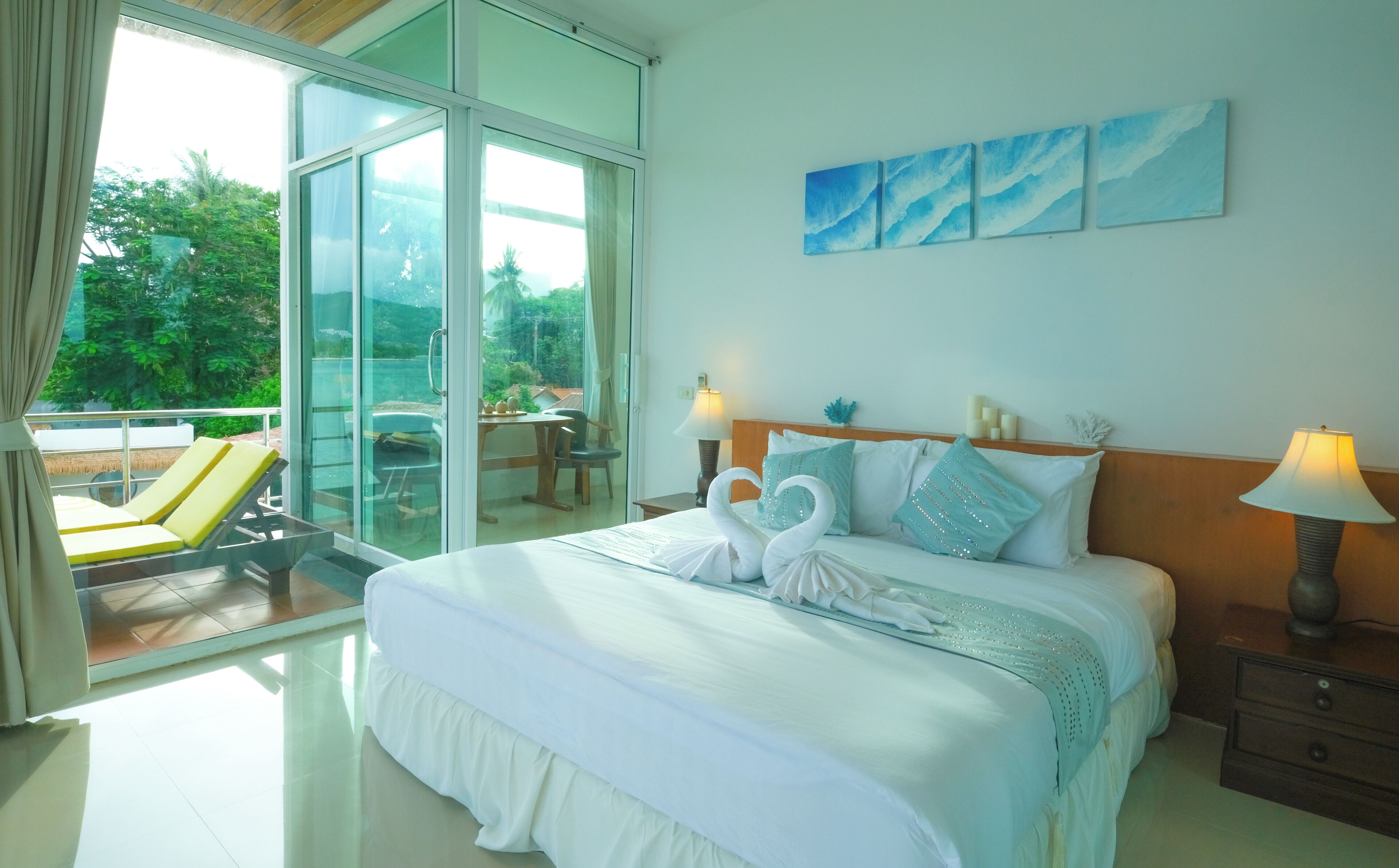 A hotel room with a bed, two lamps, a nightstand, a glass door leading to a balcony with a chair and table, and a view of greenery outside.