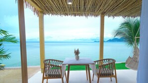 A covered outdoor seating area with a table and chairs, overlooking a body of water and green lawn. - Smile Samui Chaweng Beach Front Resort (Koh Samui)