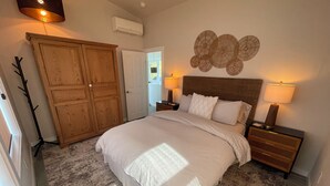 A bedroom with a large bed, wooden wardrobe, two bedside tables with lamps, and a wall decoration. - Ocean Views, Beach Path, Private + Pickleball + Nature Baths (Grayland)
