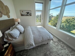 Une chambre avec une grande fenêtre, un lit avec une literie blanche, une tête de lit, une lampe fixée au mur et un tableau encadré.