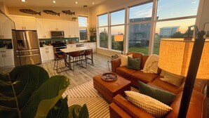 A modern kitchen with stainless steel appliances, a dining area with a table and chairs, a living room with a sofa and a coffee table, and large windows offering a view of the outside. - Ocean Views, Beach Path, Private + Pickleball + Nature Baths (Grayland)