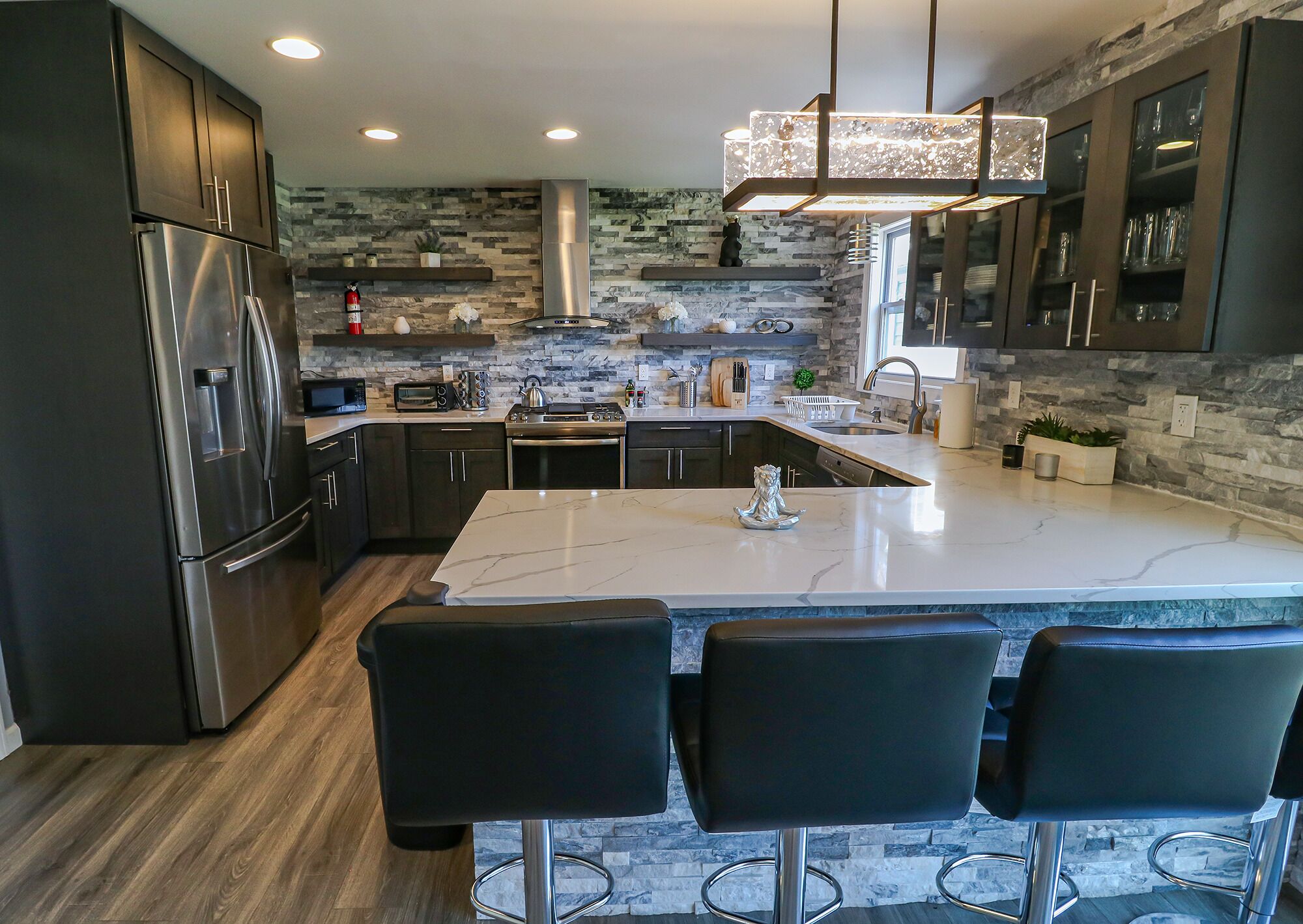 A modern kitchen with dark cabinetry, a stainless steel refrigerator, a built-in oven, a microwave, and a large island with bar stools.