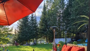 An outdoor patio with a wooden table and red cushions, surrounded by a wooden deck and a view of a forested area. - Lakefront tranquil chalet with spa near all outdoor activities (Val-Morin)