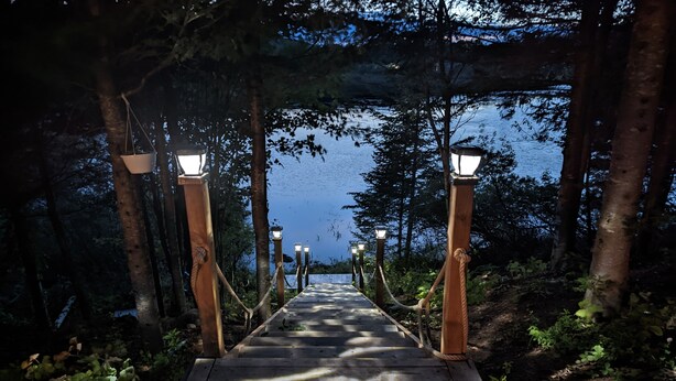 A wooden pathway leading to a lake, flanked by trees and illuminated by lanterns. - Lakefront tranquil chalet with spa near all outdoor activities (Val-Morin)