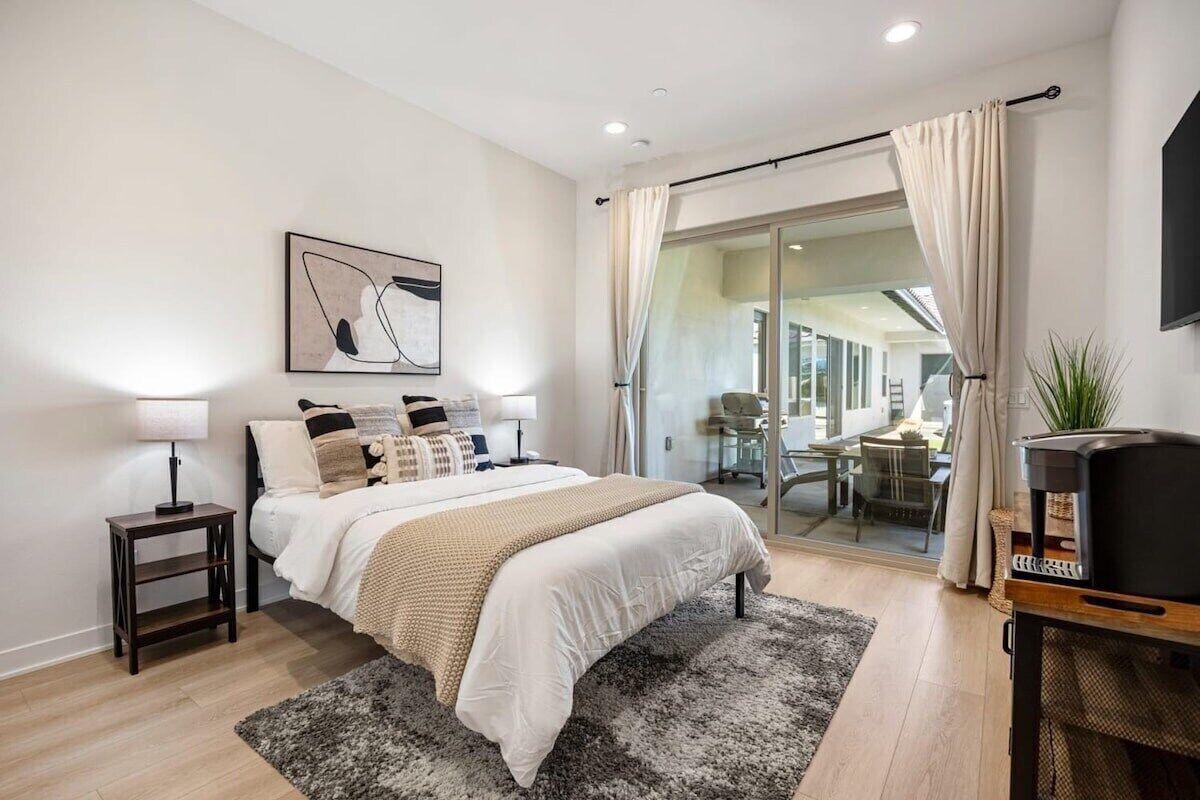 A modern bedroom with a large bed, a bedside table, a TV stand, and a view through a glass door.