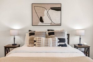 Una cama bien hecha con almohadas a rayas, dos lámparas de noche y una obra de arte abstracta en la pared.