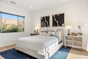 A modern bedroom with a large bed, two bedside tables, a nightstand, a console table, and a window with a view of greenery.