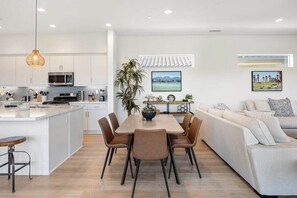 Una cocina moderna con armarios blancos, un comedor con mesa y sillas, y una sala de estar con sofá y cojines.