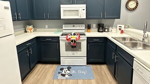 Una cocina moderna con armarios azul oscuro, un microondas blanco y una estufa. Hay un reloj en la pared y un tapete con un personaje de dibujos animados en el suelo.