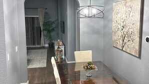 Un comedor con una mesa de madera, sillas blancas, una araña y un cuadro colgado en la pared.