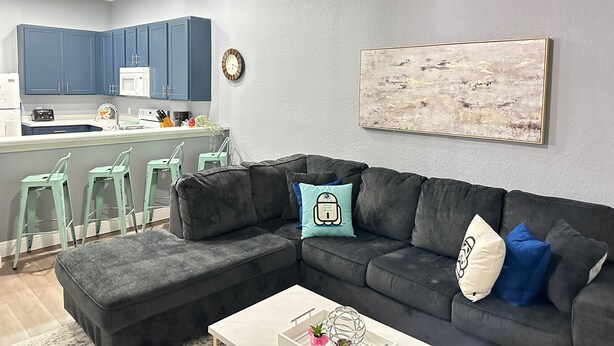 Una sala de estar moderna con un sofá seccional gris, una mesa de centro y una cocina con armarios azules y una encimera blanca.