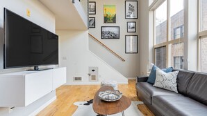 Uma sala de estar moderna com uma TV de tela plana, um sofá com almofadas estampadas, uma mesa de centro de madeira e uma escada decorada com quadros.