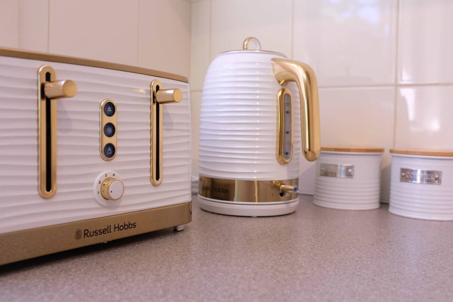 Une cuisine avec un appareil électroménager blanc et doré, une bouilloire blanche et deux récipients blancs avec des couvercles dorés.
