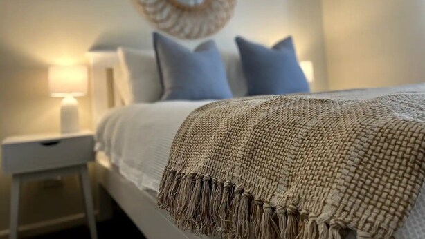Una cama bien hecha con una manta tejida y almohadas, una lámpara de noche y un espejo montado en la pared.