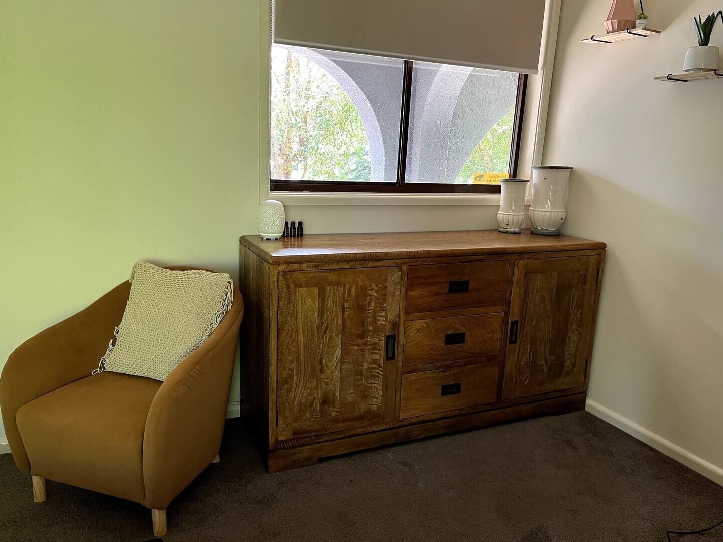 Une pièce comprenant un fauteuil en cuir marron, une commode en bois avec des tiroirs, une fenêtre donnant sur des arbres et une étagère ornée d’objets décoratifs.