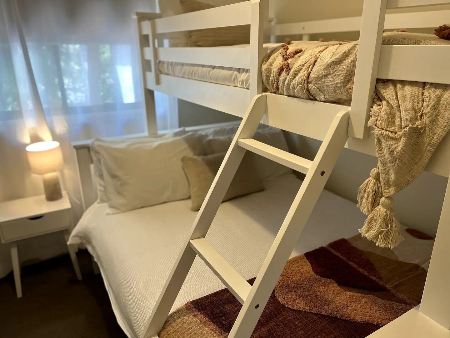 A bunk bed with a ladder, a bedside table with a lamp, and a view of the outdoors through a window.