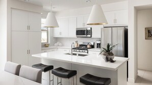 Une cuisine moderne avec des meubles blancs, un îlot central doté de tabourets de bar, des appareils en acier inoxydable et un éclairage suspendu.