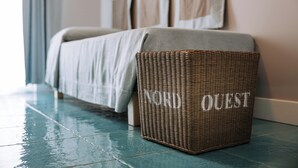 Un reposapiés de mimbre con la palabra “QUEST”, sobre un suelo de baldosas azul verdoso, junto a una cama con una manta color beige.