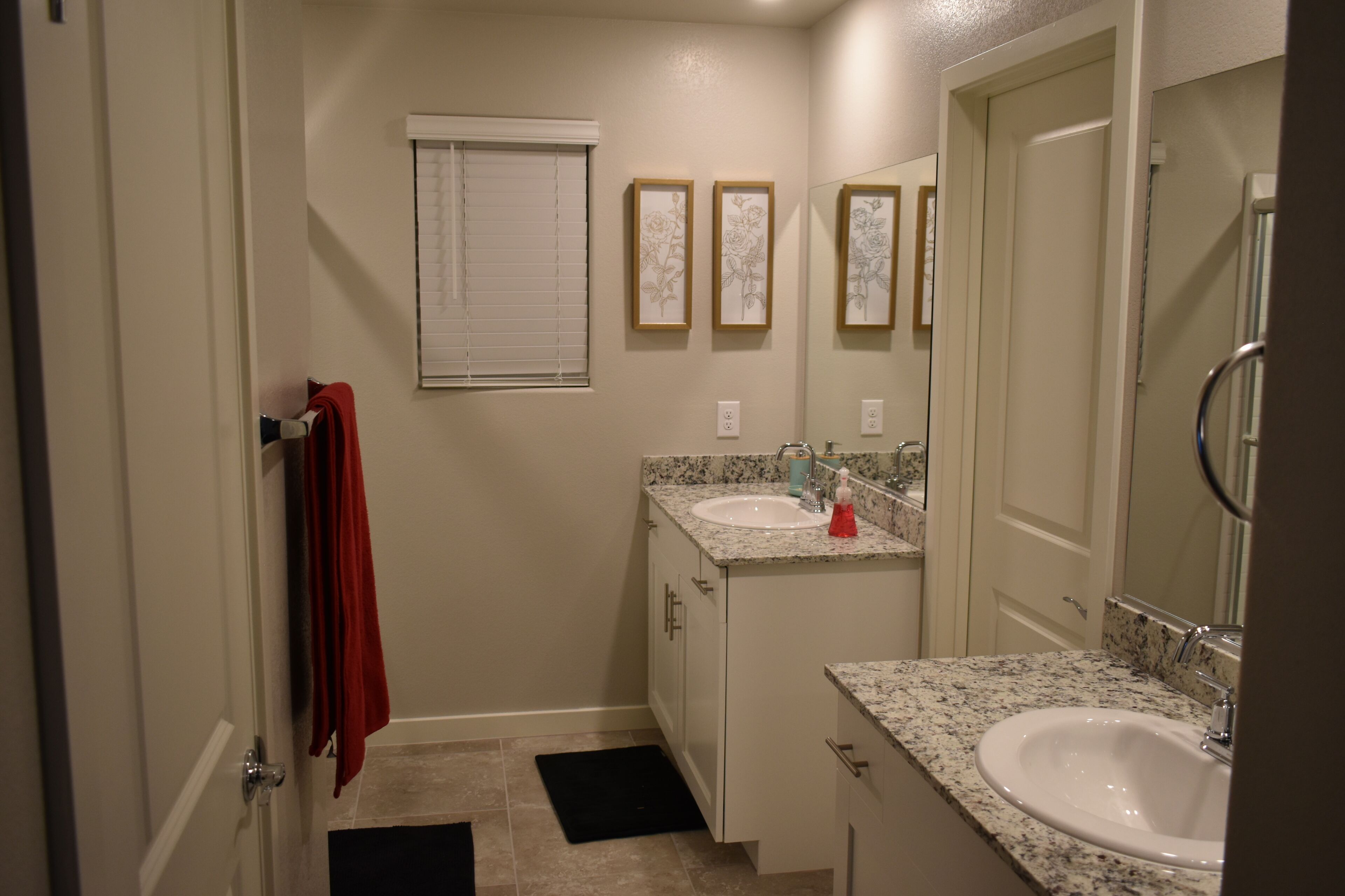 A small bathroom with two sinks, a window with blinds, and a mirror.