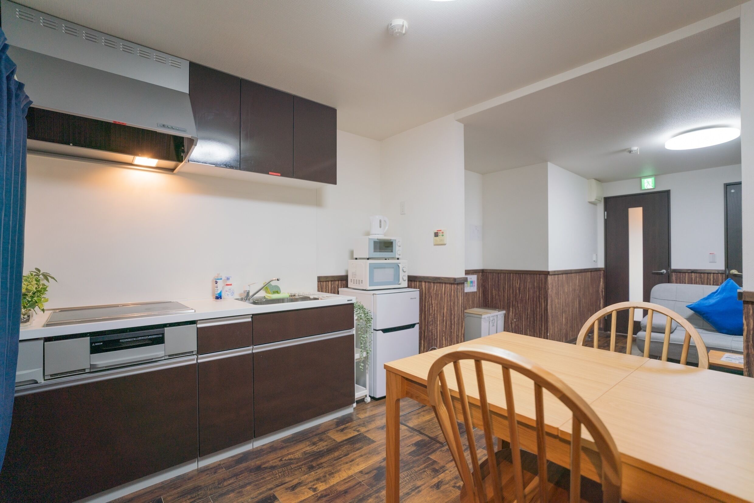 A compact kitchen with a microwave, refrigerator, and dishwasher. A wooden dining table with chairs is in the foreground.