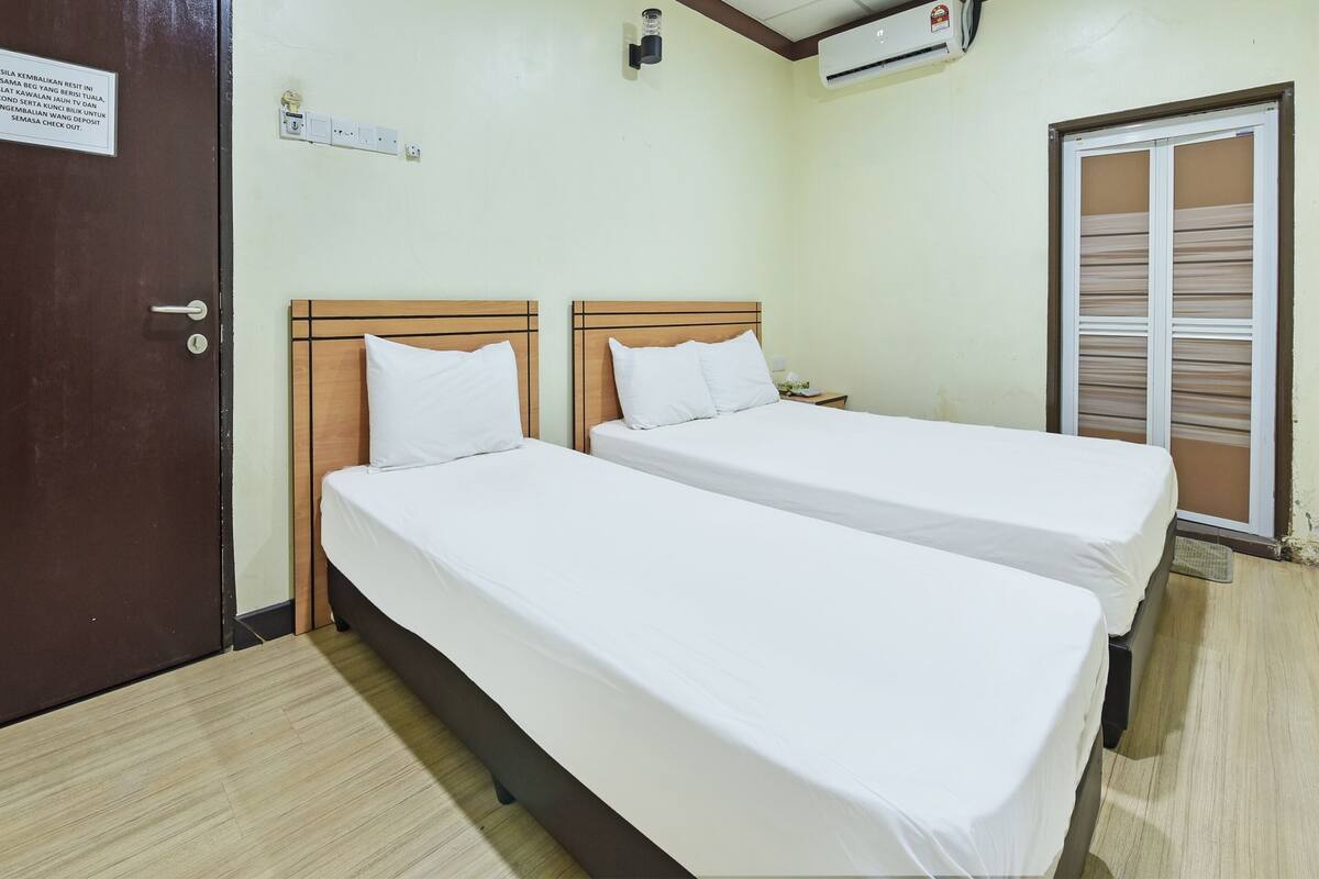 Hotelkamer met twee eenpersoonsbedden, een houten hoofdbord, een klein nachtkastje met een lamp, een wandairconditioner en een deur met een bordje.
