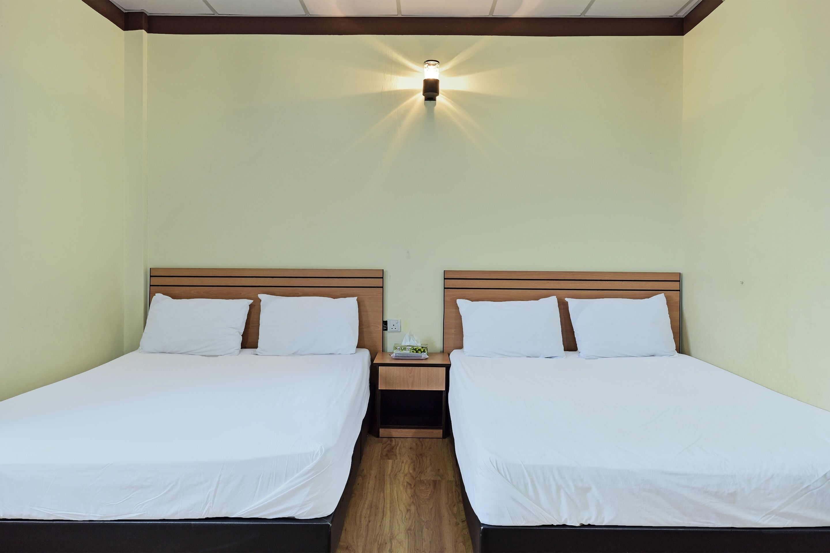 Quarto de hotel com duas camas de solteiro, cabeceiras de madeira, mesa de cabeceira com abajur e luz no teto.