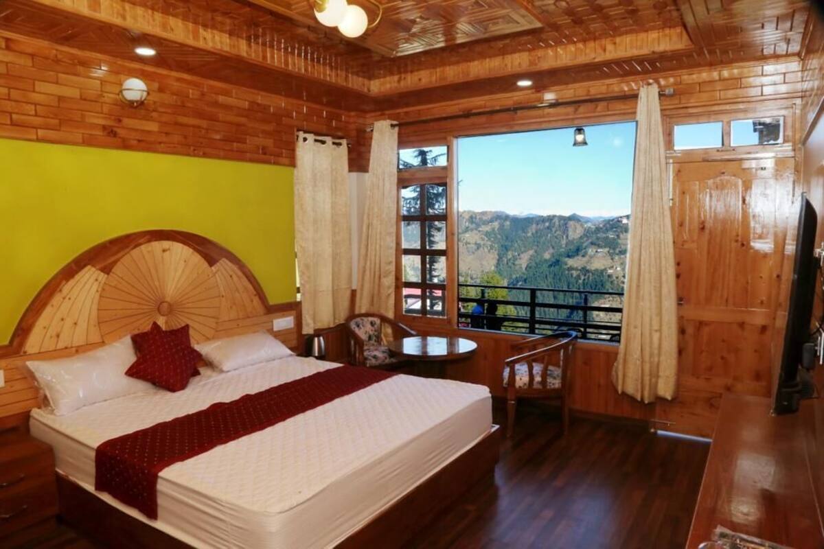 Ein Schlafzimmer mit Holzboden, einem großen Bett, einem kleinen Tisch und einem Stuhl. Ein Fenster bietet Blick auf die Berge.