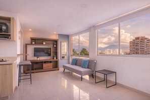 A modern living room with a flat-screen TV, wooden entertainment unit, grey sofa, and a city view through large windows.