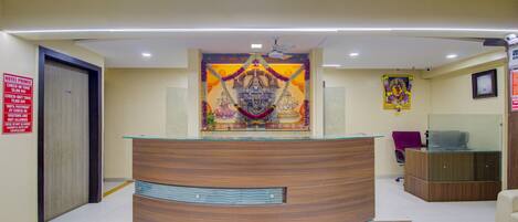 Un mostrador de recepción con detalles de madera y una superficie de vidrio, flanqueado por una pared con un mural religioso y un ventilador de techo.