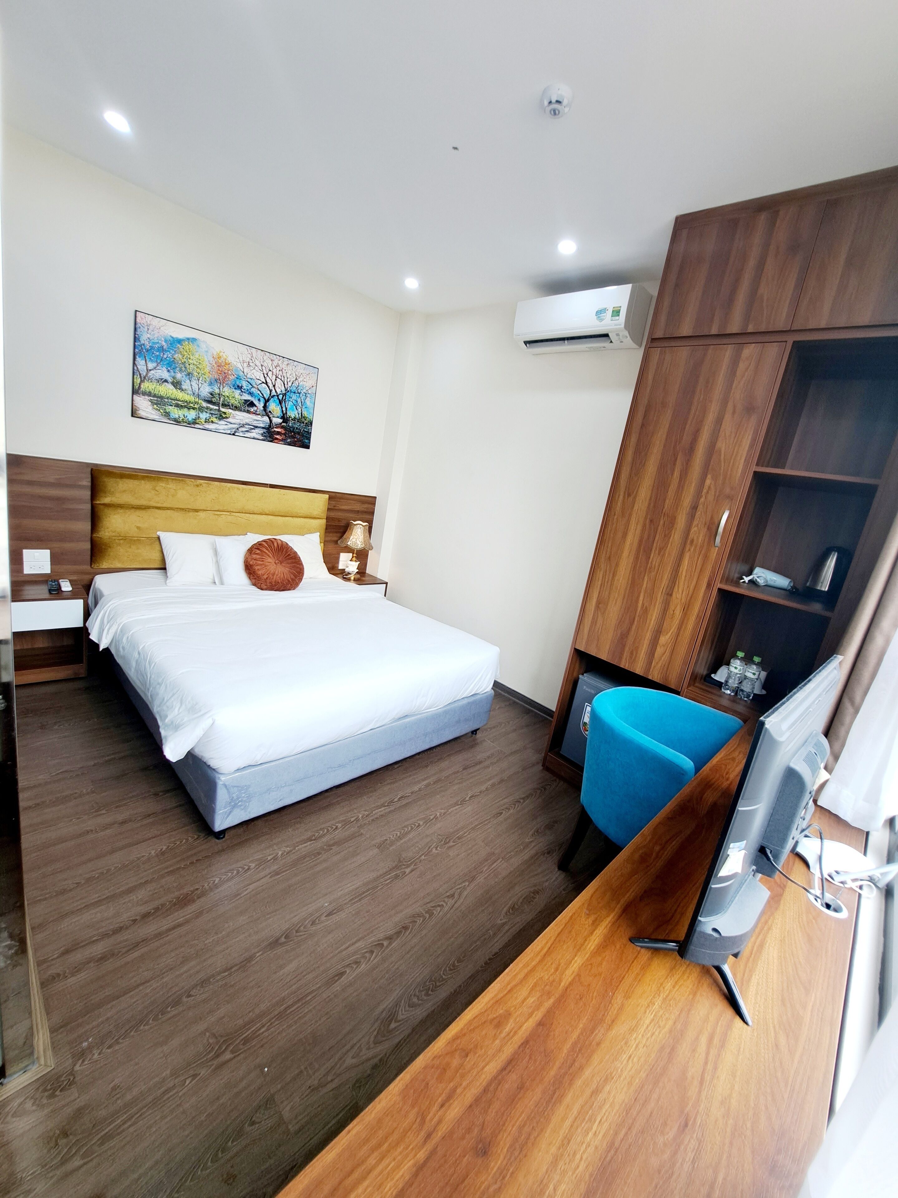 A hotel room with a bed, a desk with a computer, a chair, a wardrobe, and an air conditioning unit.