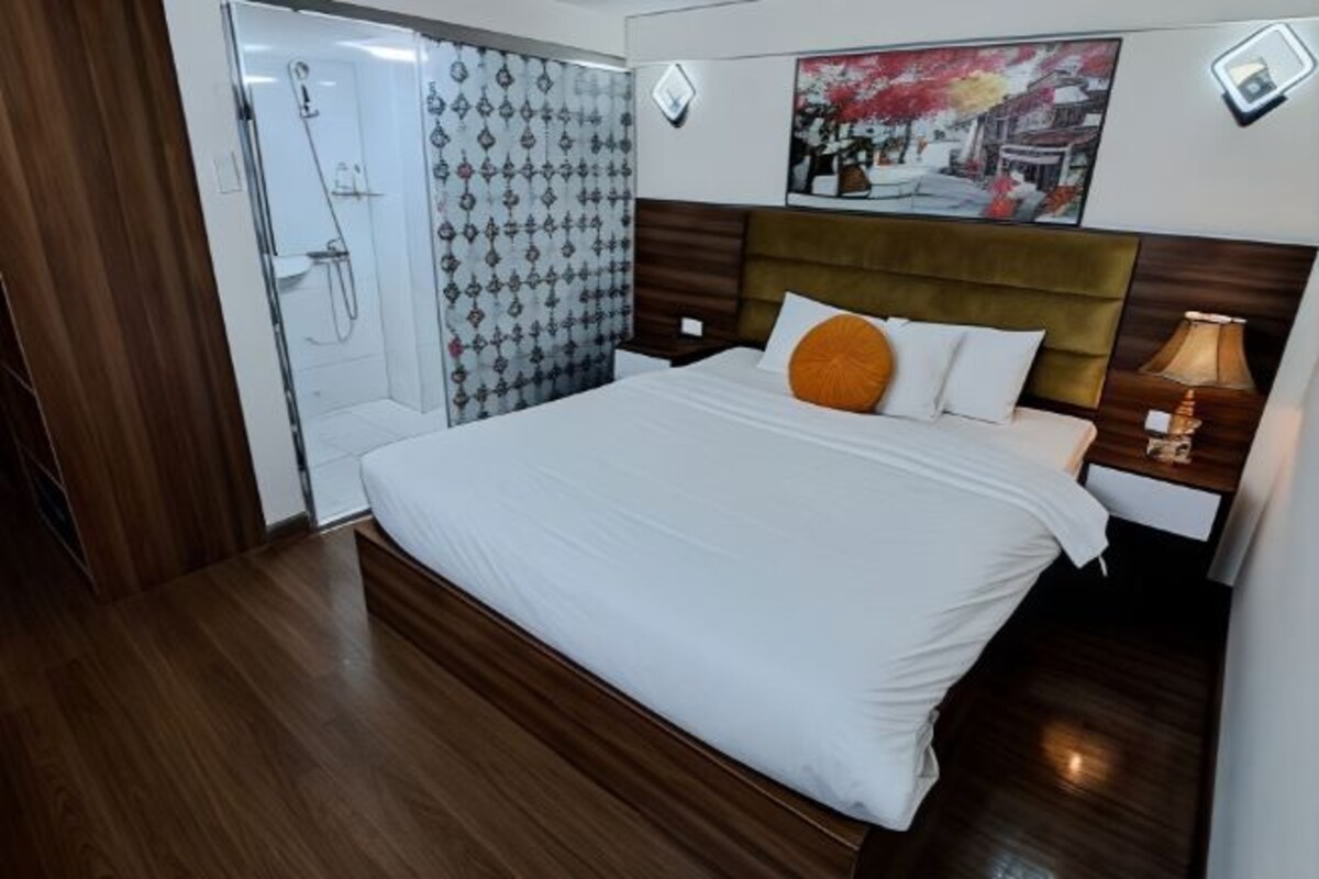 Habitación de hotel con piso de madera, cama con ropa blanca y almohada naranja, mesita de noche con lámpara y zona de ducha con cortinas estampadas.