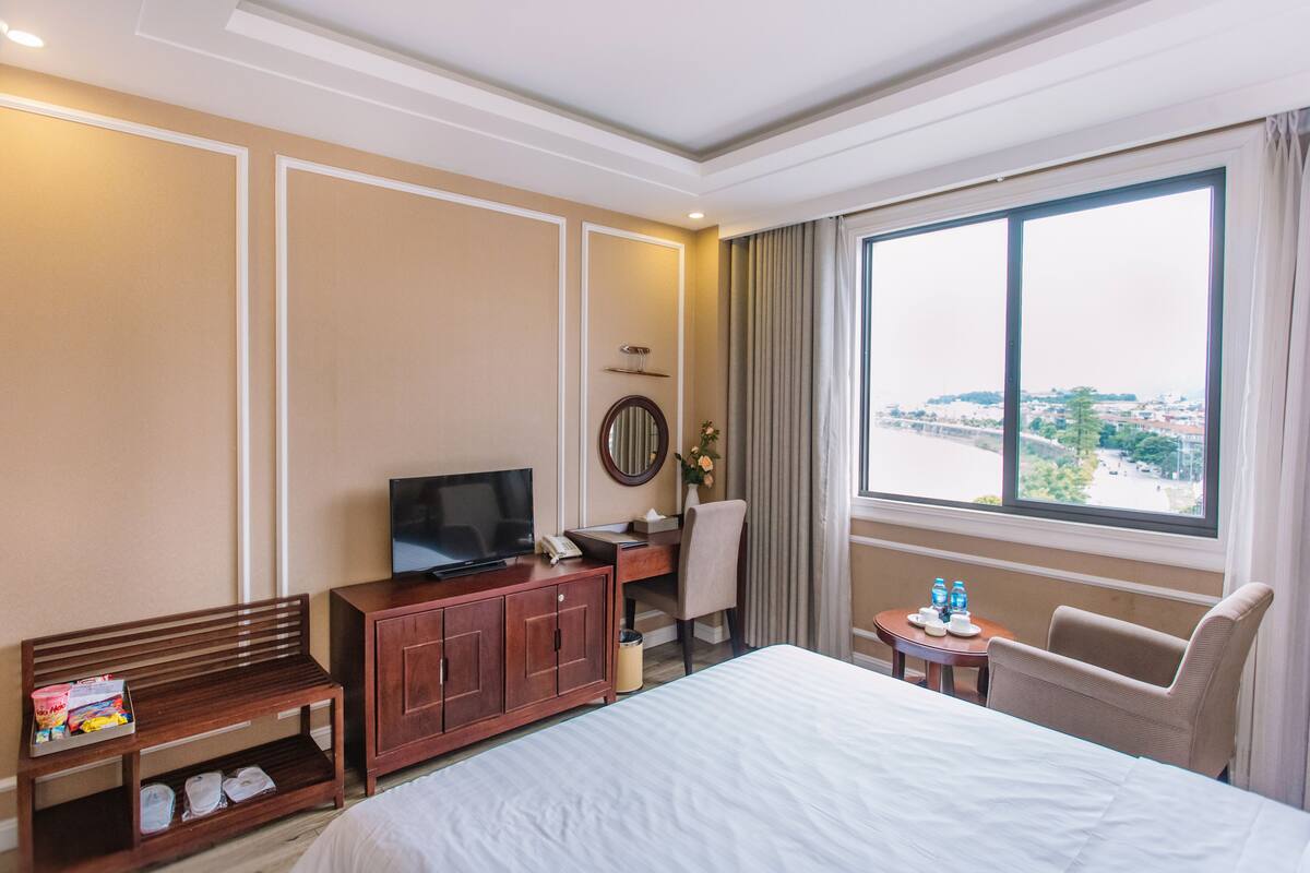 Habitación de hotel con una cama grande, un escritorio con televisor, una silla, una mesita con botellas de agua y vistas a la ciudad a través de un ventanal amplio.