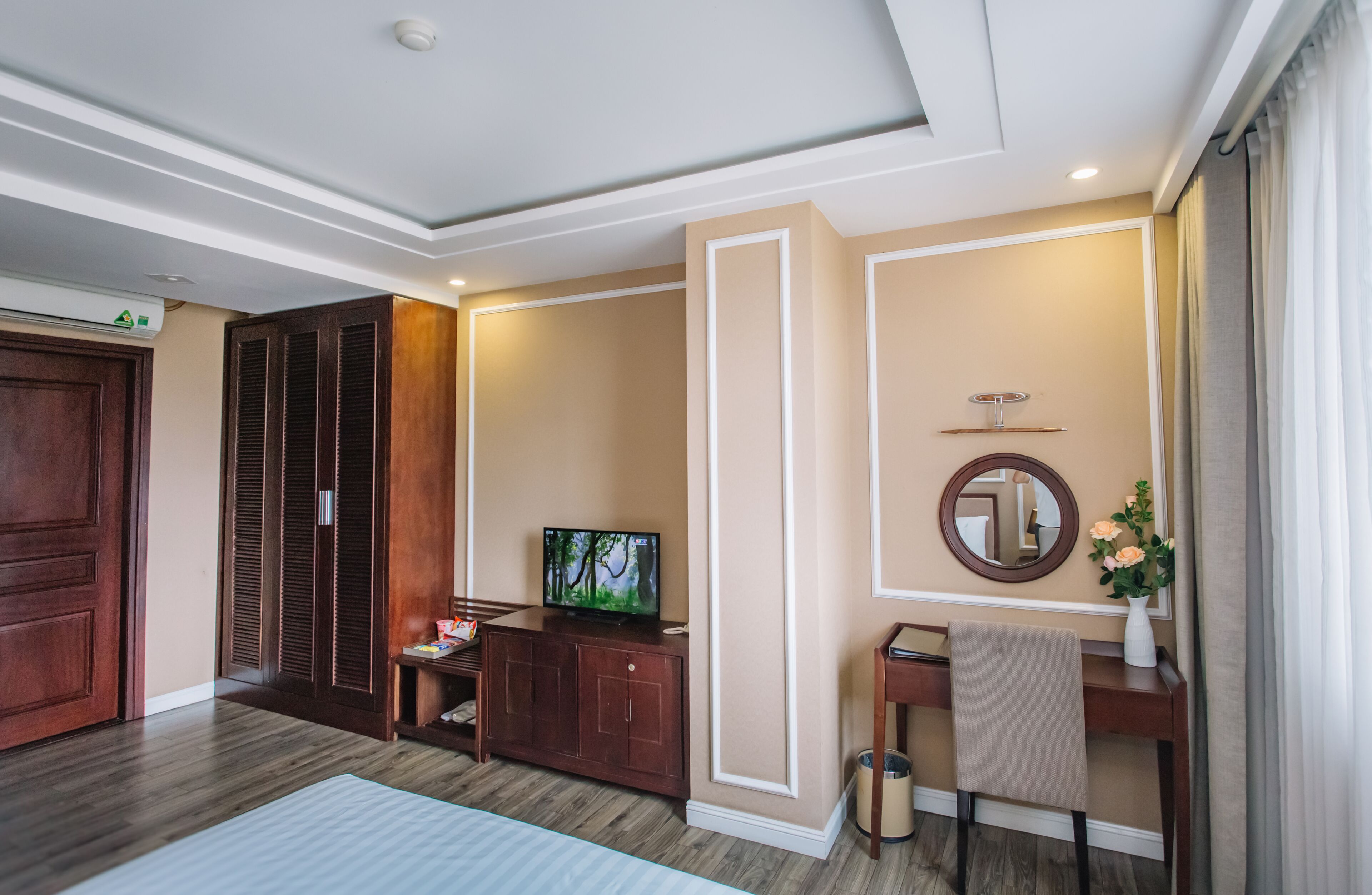 Une chambre d’hôtel avec une armoire en bois, un téléviseur posé sur un meuble, un bureau avec une chaise, un miroir et un vase avec des fleurs.