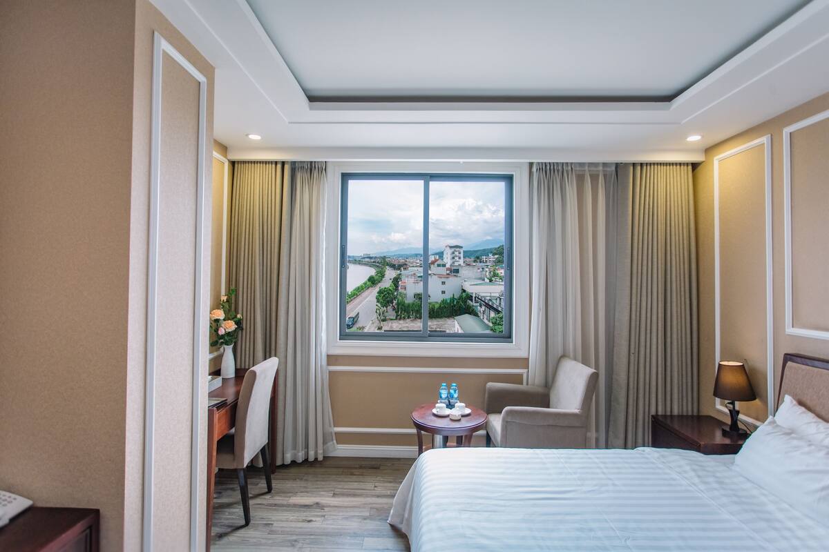 Una habitación de hotel con un ventanal grande, una cama, un escritorio, una silla y una mesita con una lámpara.