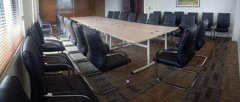 Un salón de conferencias con una larga mesa y varias sillas, piso alfombrado y una ventana con persianas.