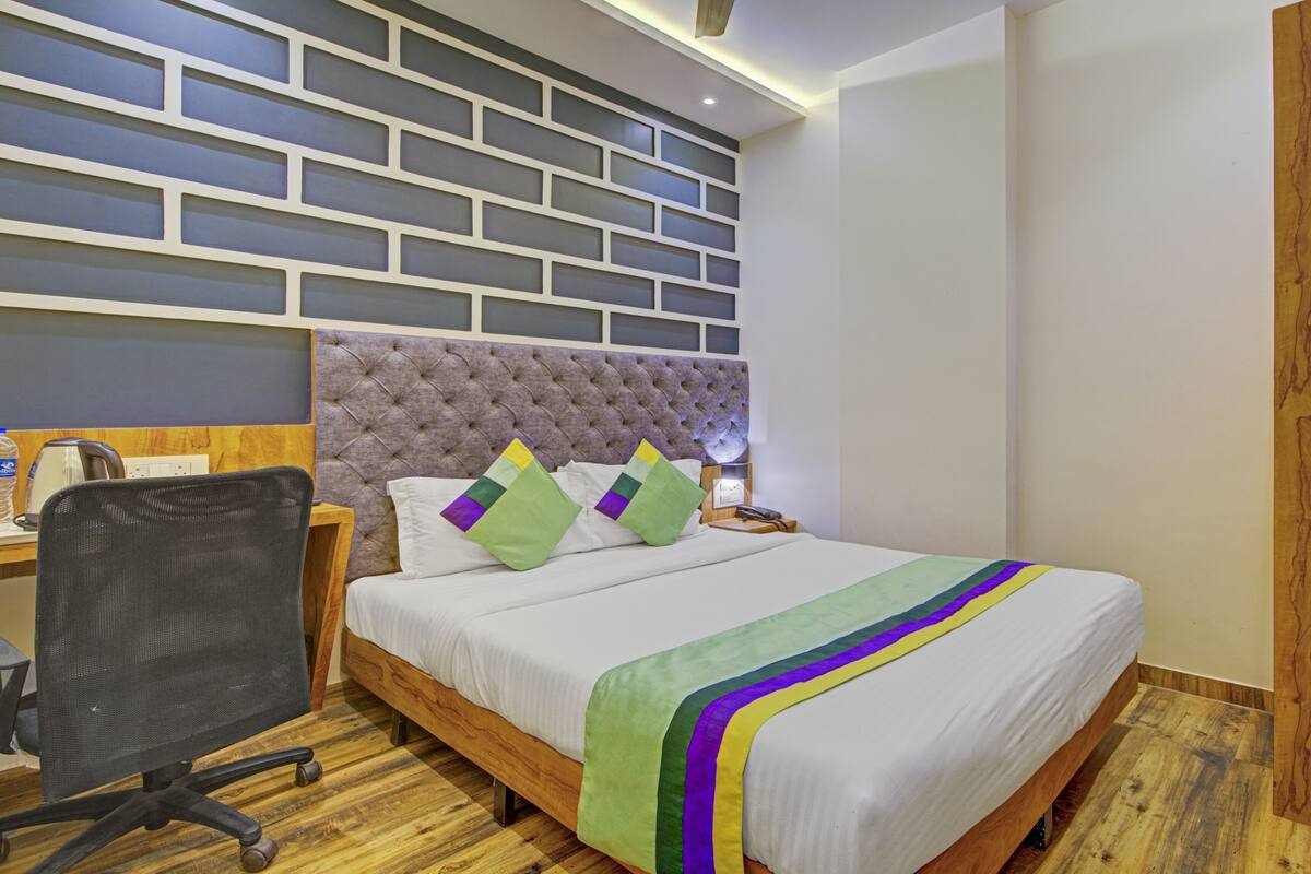 A hotel room with a wooden floor, a bed with a green and purple striped bedspread, a tufted headboard, and a desk with a chair.