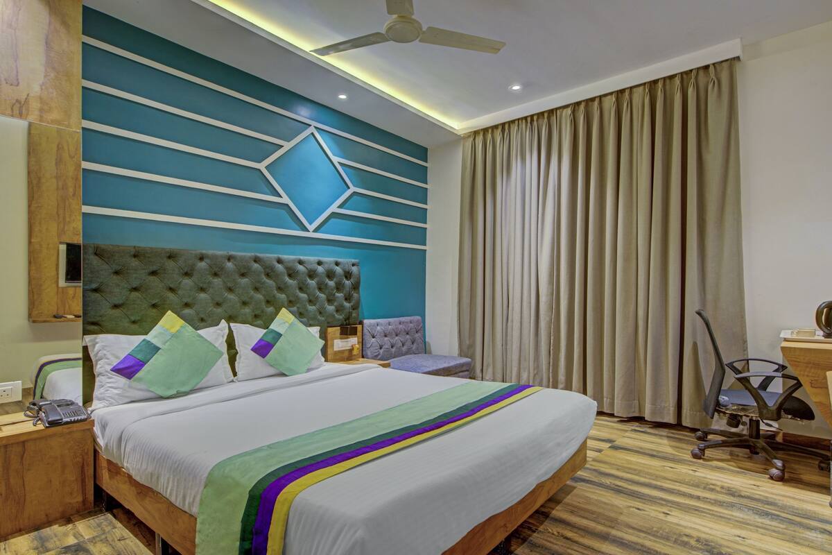A hotel room with a large bed, a tufted headboard, a desk with a chair, a ceiling fan, and a window with curtains.