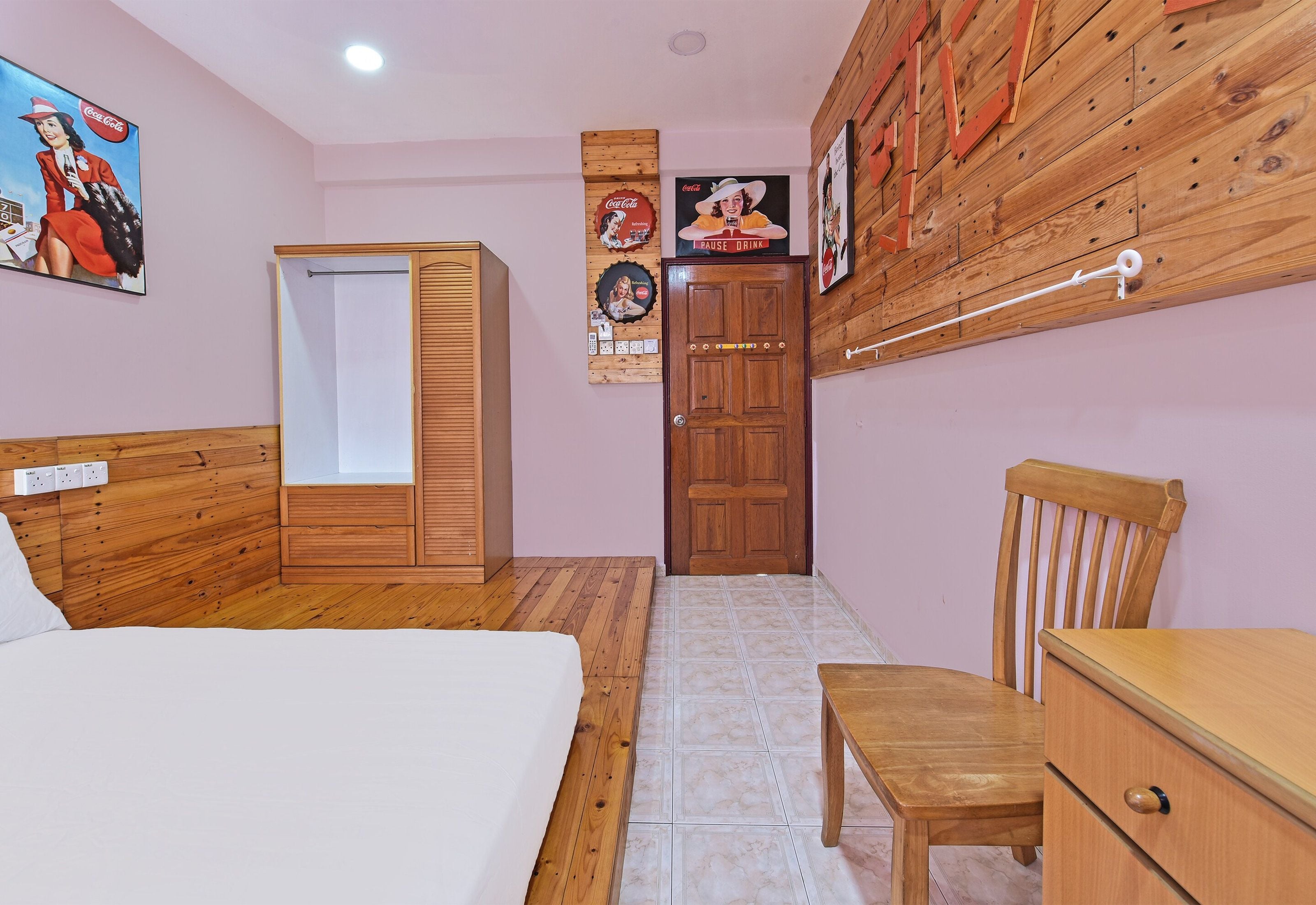 A bedroom with a bed, a wooden wardrobe, a desk, and a chair. There are posters on the wall and a door with a handle.