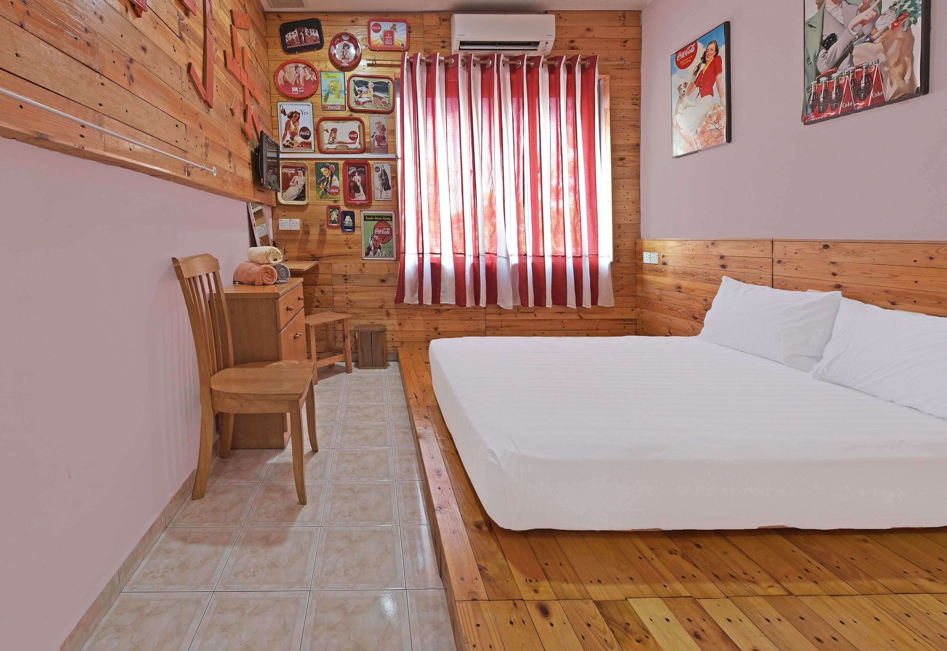 Un dormitorio con cabecera de madera, cama, una silla, una mesita y una pared decorada con recuerdos antiguos de Coca-Cola.