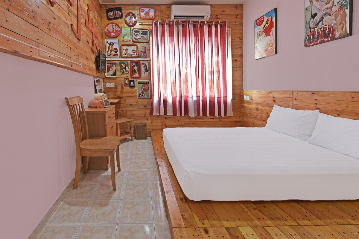 Een slaapkamer met een houten hoofdeinde, een bed, een stoel, een klein tafeltje en een muur versierd met verzamelobjecten van Coca-Cola uit vervlogen tijden.