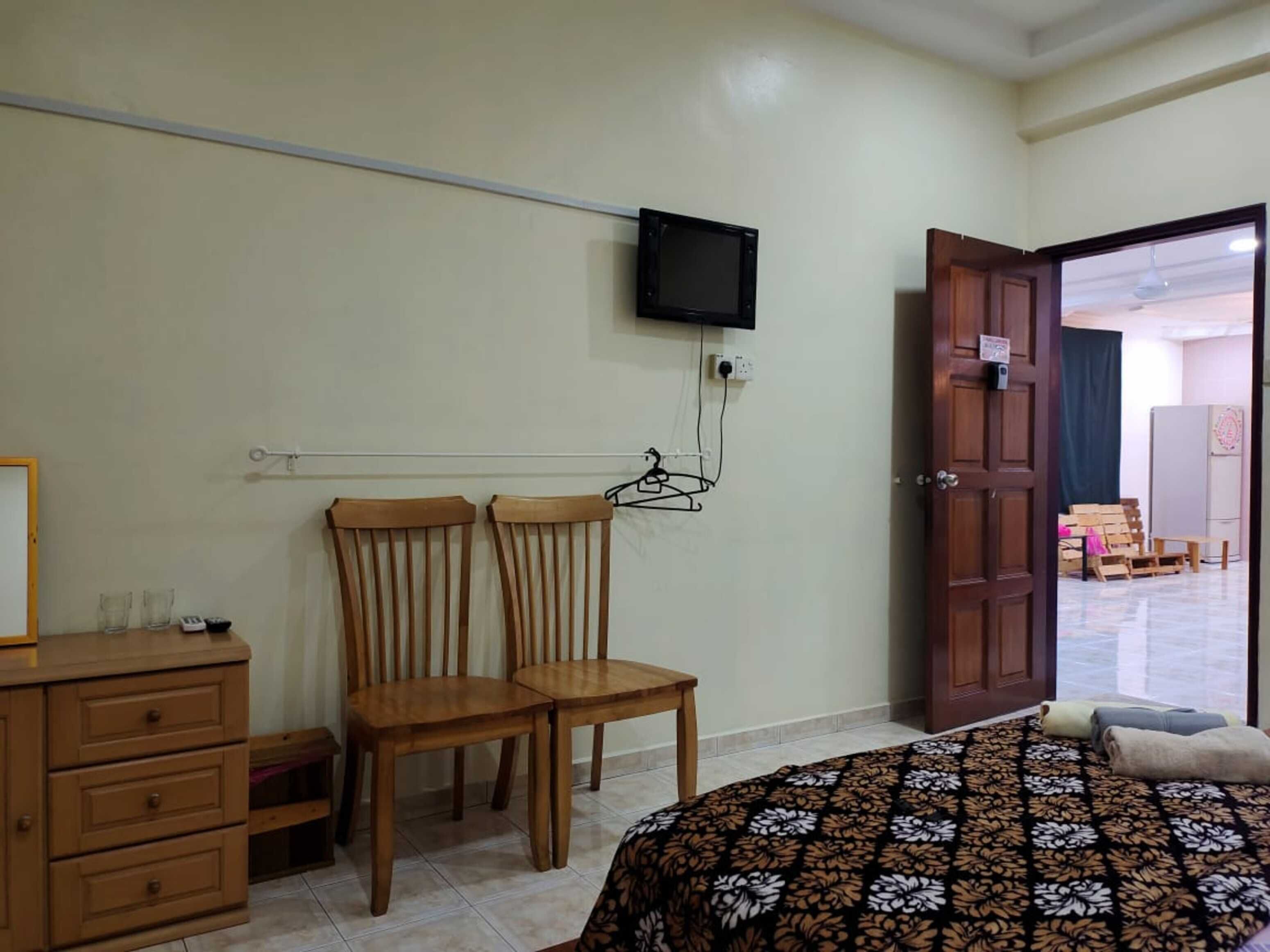 Un dormitorio con cama, dos sillas de madera, cómoda, televisor montado en la pared y una puerta abierta que da a otra habitación.