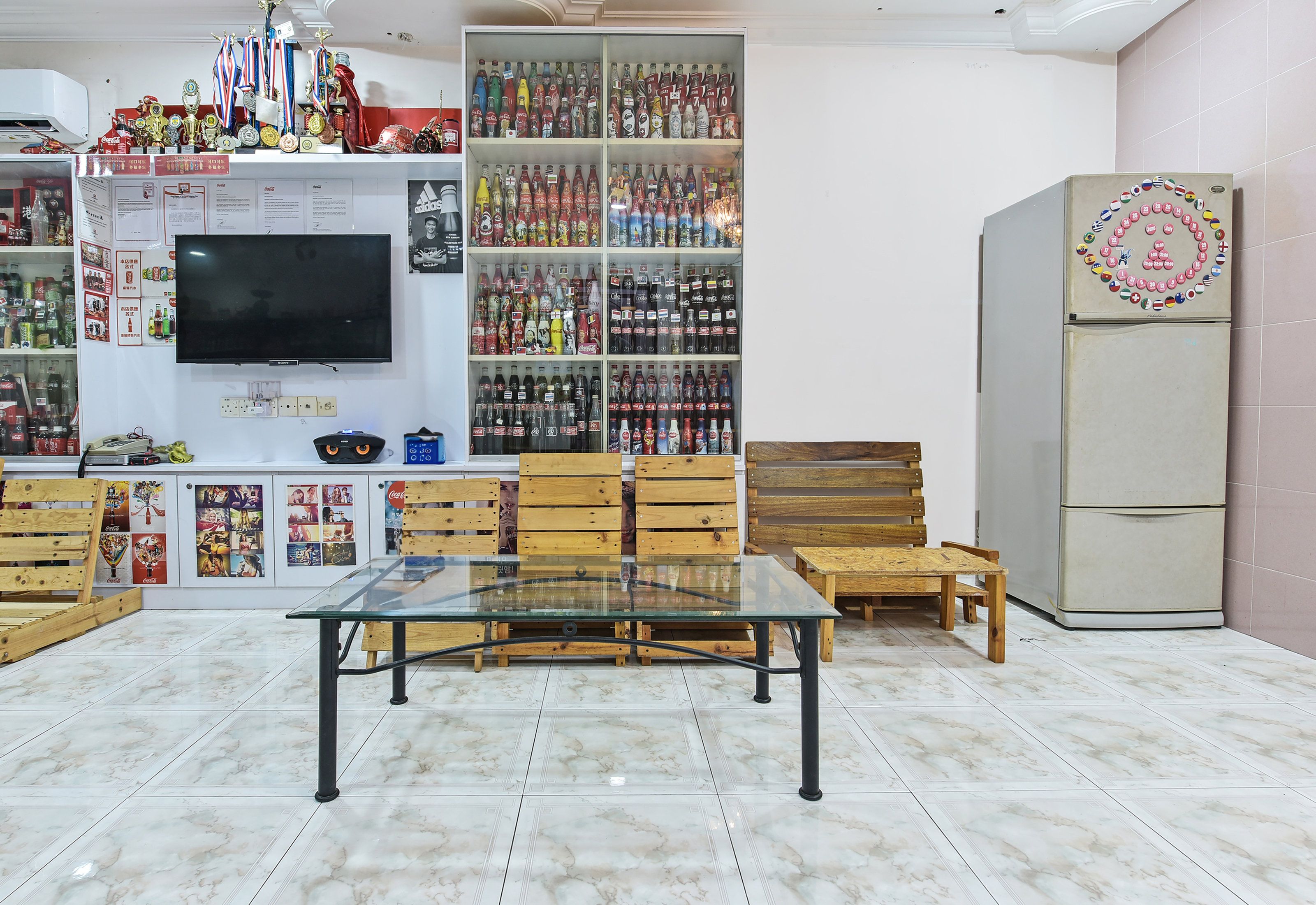 A room with a glass-top table, wooden chairs, a television, shelves with various items, and a refrigerator.