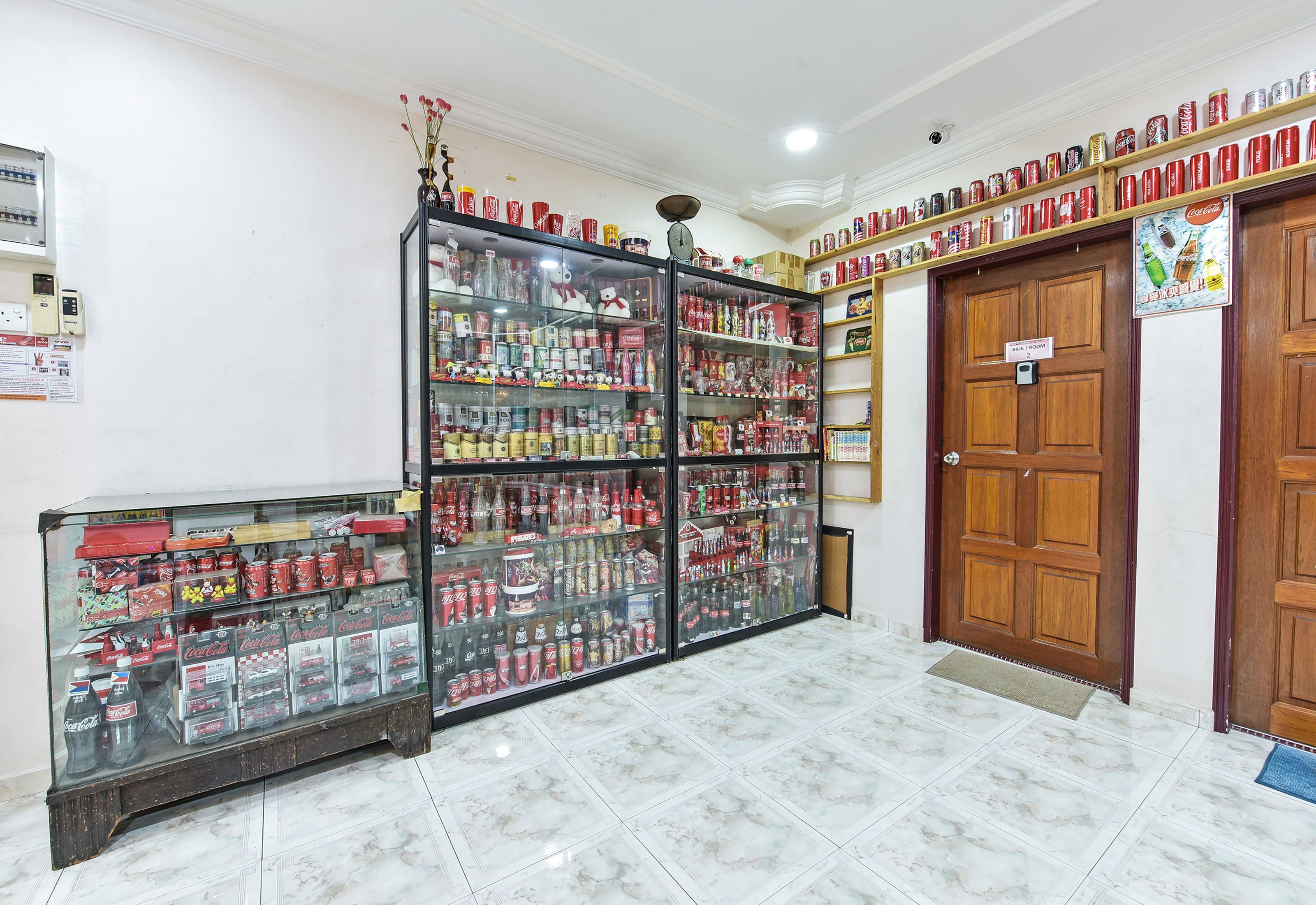 Um quarto com uma vitrine de vidro cheia de conservas e bebidas, uma porta de madeira com uma placa, e um chão de azulejos.