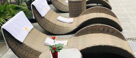 Una hilera de sillones de mimbre con toallas blancas, dispuestas en un patio empedrado con una piscina al fondo.