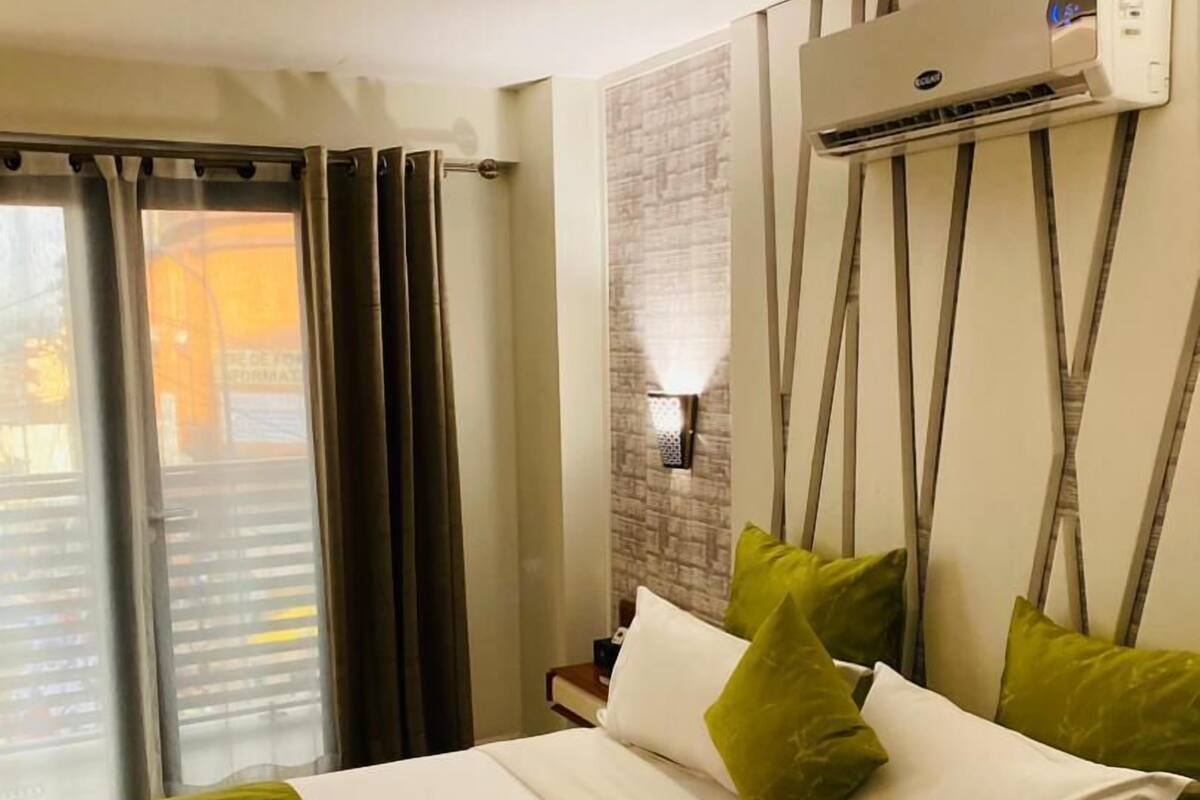 A hotel room with a bed, curtains, an air conditioning unit, and a wall-mounted light fixture.