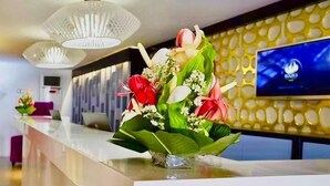 A reception area with a white reception desk, a large TV screen displaying a logo, and a floral arrangement on the desk. - Boun's Hotel Resort & Spa (Yaoundé)