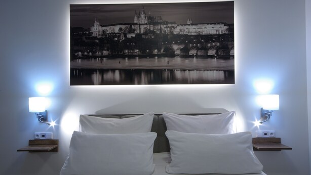 A hotel room with a bed, two bedside tables, and a wall-mounted picture of a cityscape. - Adeba Plus (Prague)