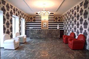 Uma área de recepção com um balcão de mármore, duas poltronas brancas, duas poltronas vermelhas e um lustre.