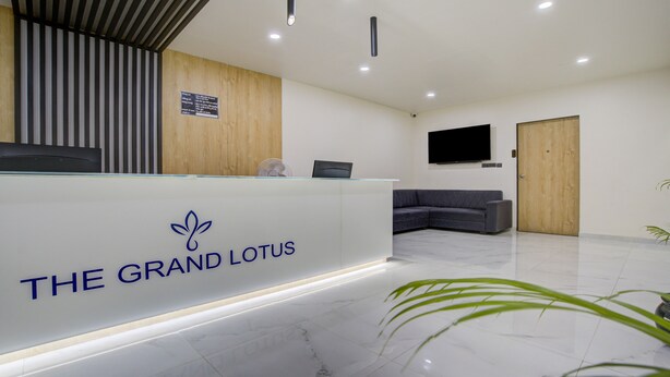 Un área de recepción con una mesa de recepción identificada como "THE GRAND LOTUS", un panel de pared de madera, un televisor de pantalla plana, un sofá gris y una puerta de madera.